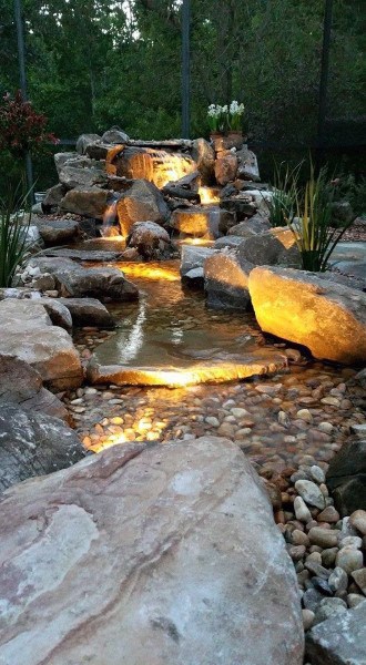 Backyard waterfall with illuminated stream, natural rocks, and a serene garden setting.