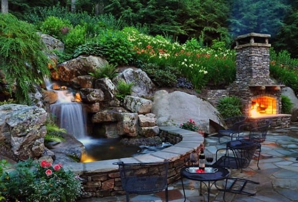 Raised backyard waterfall pond with natural stones, lush greenery, and an outdoor fireplace.