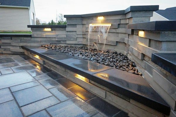 Modern backyard waterfall with sleek stone design, black pebbles, and integrated lighting.