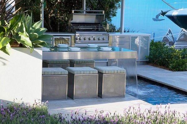 Outdoor kitchen with stainless steel countertop featuring a built-in waterfall and modern seating.