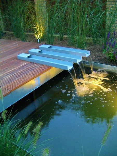 Modern pond with triple stainless steel waterfall spouts and soft evening lighting.
