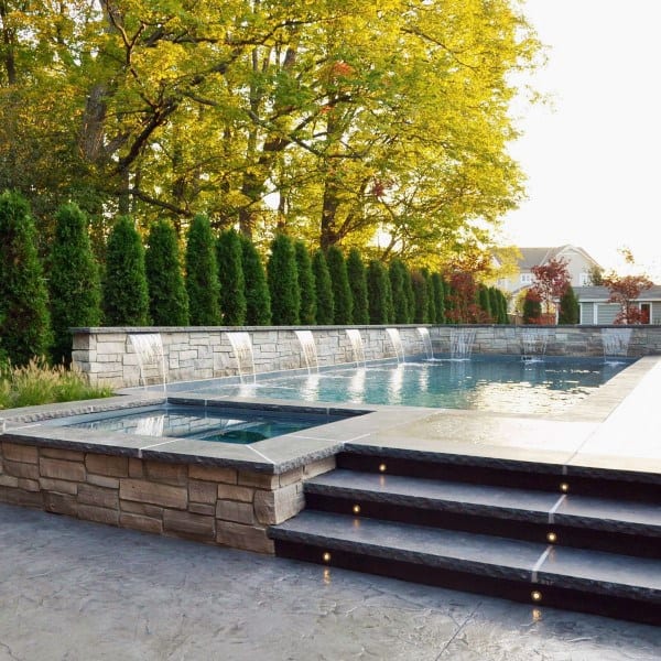 Elegant backyard pool with multiple waterfalls, stone accents, and a landscaped hedge backdrop.