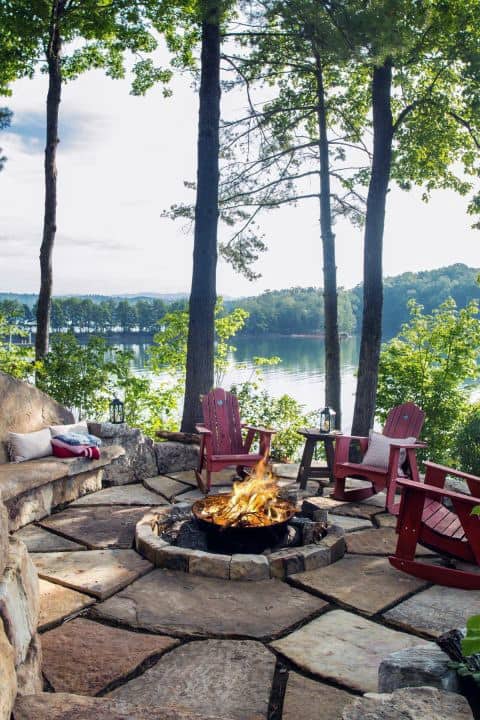 Cozy lakeside fire pit with red chairs, surrounded by trees and stone seating, overlooking a serene lake