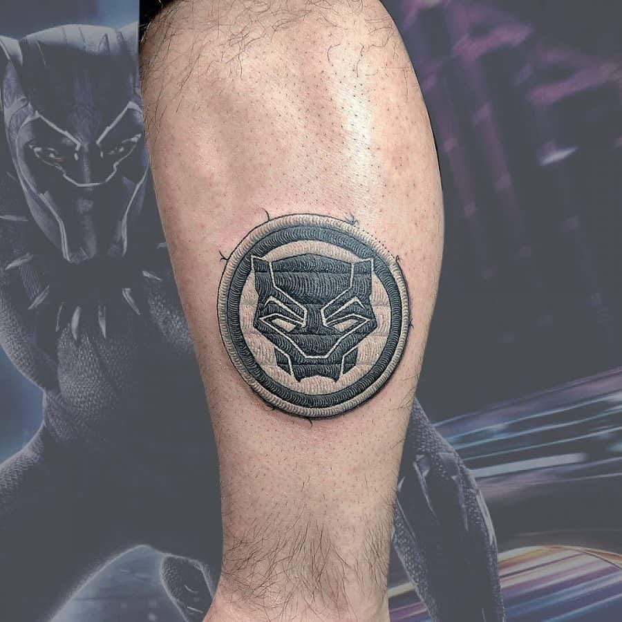 Black and grey embroidery tattoo of a superhero emblem, giving the appearance of a stitched patch on the skin.