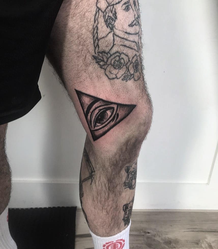 Tattoo of an all-seeing eye inside a shaded triangle, inked in black and grey realism on the upper leg.