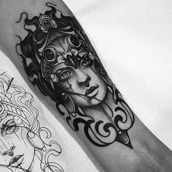 Surreal Athena tattoo with expressive eyes, ornate helmet, and bold ornamental patterns on the inner forearm in black and grey style
