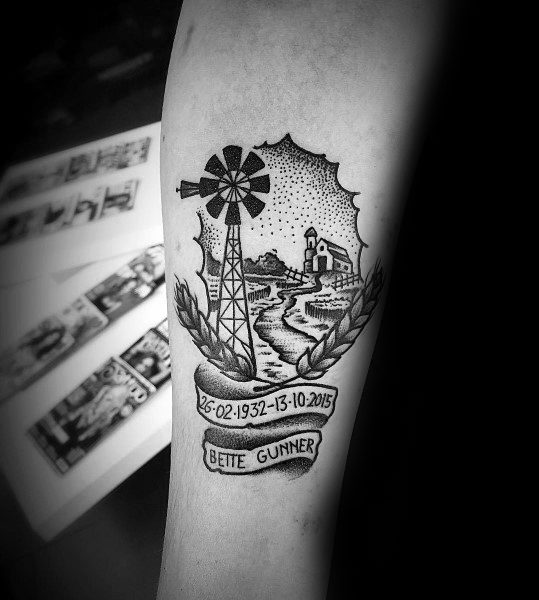 Black and gray farming tribute tattoo featuring a windmill, barn, fields, and a memorial banner with a name and dates.