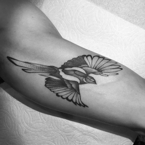 Black and gray magpie tattoo on the inner arm, featuring the bird in mid-flight with detailed feathers and smooth shading for a dynamic look.