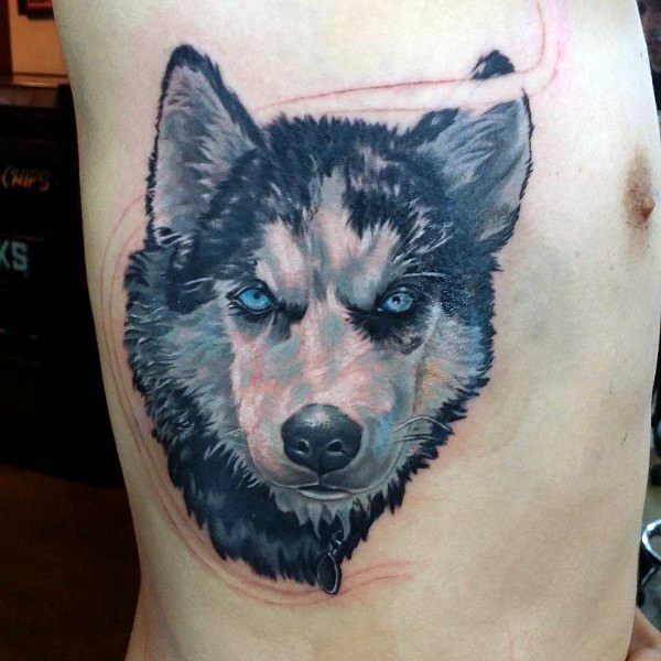 Realistic Siberian Husky tattoo on a man's ribs, featuring piercing blue eyes, detailed fur shading, and a fierce expression for a bold design.