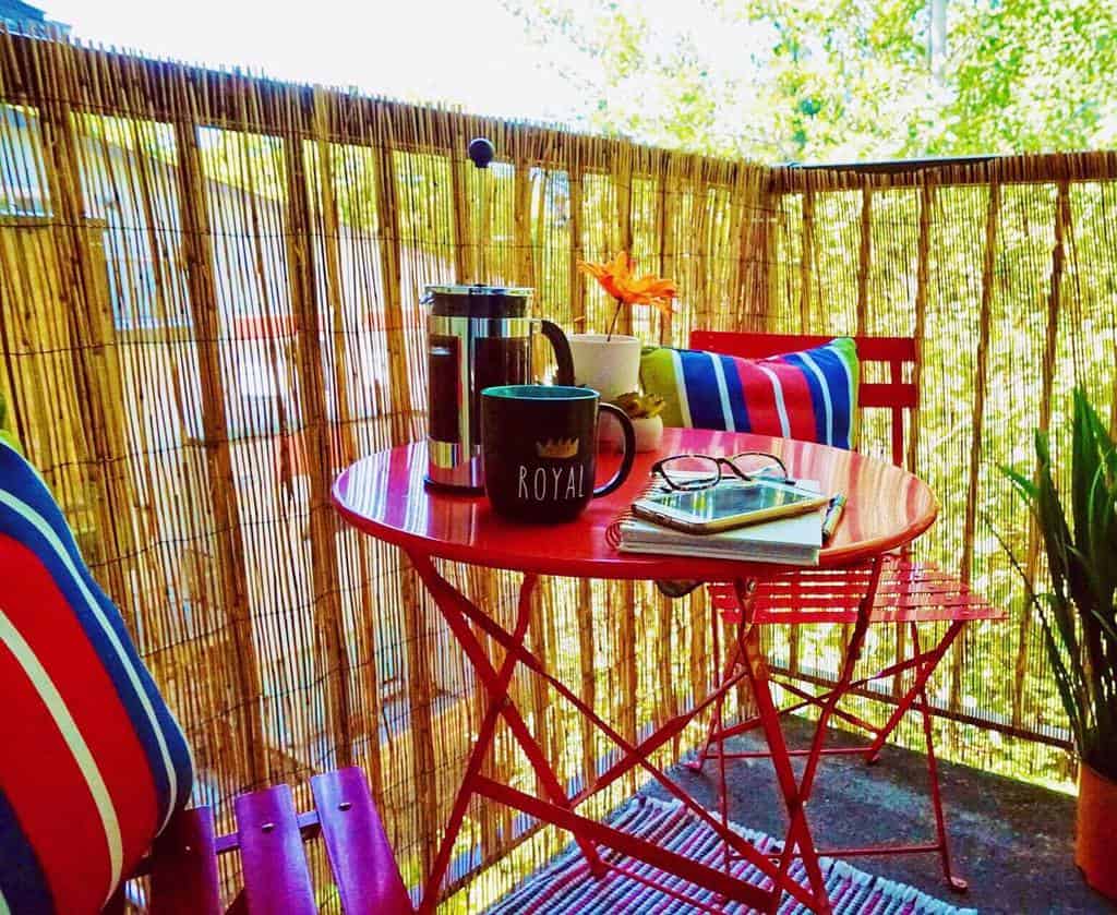 Cozy balcony with red table, coffee mugs, books, and colorful pillows on a bamboo privacy screen.