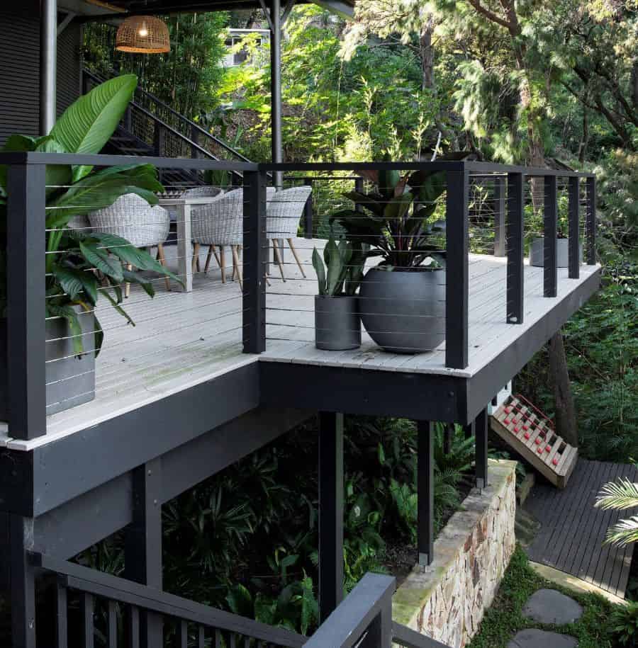 Elevated wooden deck with modern railing, potted plants, and wicker chairs amidst lush greenery. Steps lead down to garden
