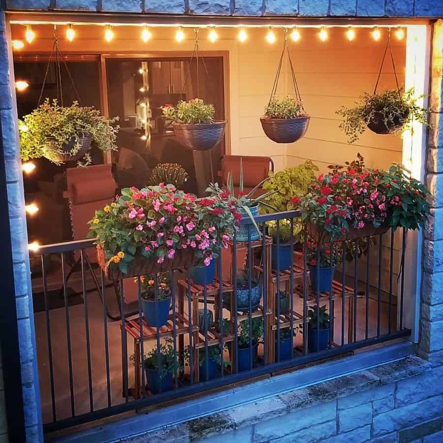 Cozy balcony with hanging plants, flowers, and string lights; two chairs face a small table with more plants, viewed from outside