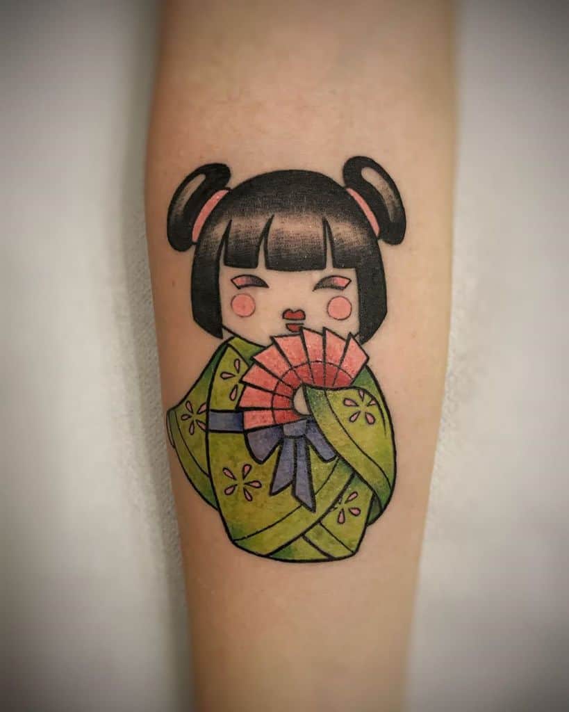 Colorful tattoo of a cute, cartoon-style Geisha with a green kimono, pink fan, and playful hairstyle, featuring a cheerful expression.
