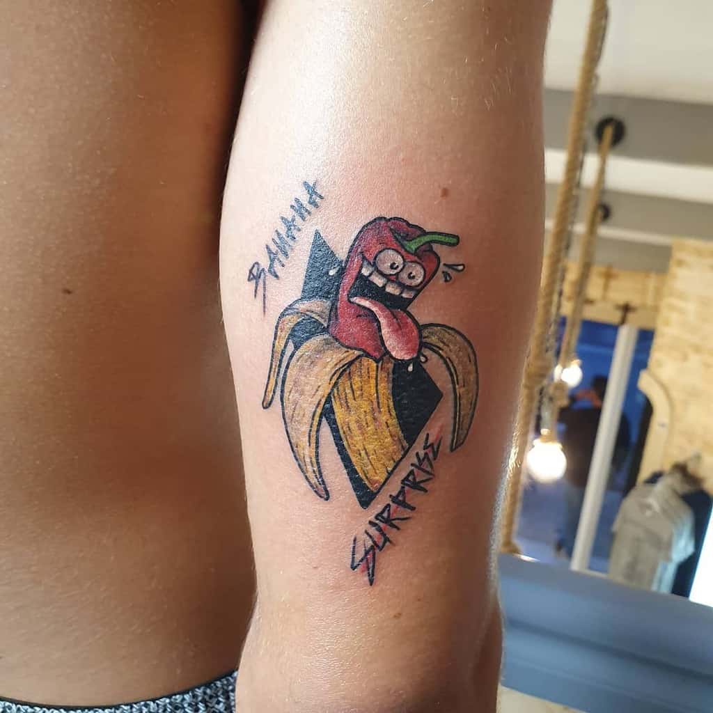 A colorful tattoo of a peeled banana revealing a shocked, cartoonish hot pepper inside, with the words 'BANANA' and 'SURPRISE' in bold lettering.