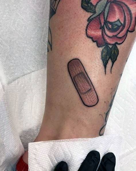 Minimalist Band-Aid tattoo with soft shading and clean outlines, placed below a rose tattoo on the lower leg.