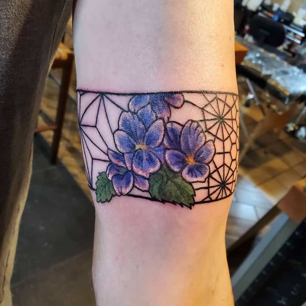 Unique armband tattoo featuring vibrant violet flower with green leaves, set against a geometric spiderweb design for a bold contrast