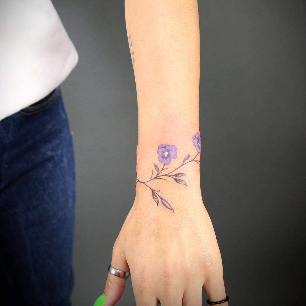 Delicate wrist tattoo of violet flowers with fine line branches, creating a minimalist and elegant floral bracelet design