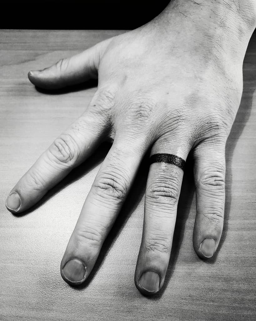 A black and white tattoo of a thick black ring on a finger, symbolizing a wedding band with a bold, minimalist design