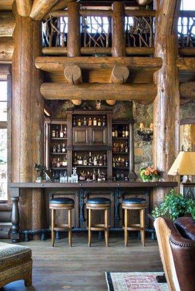 Rustic log cabin home bar with wooden stools, dark wood shelving, and stone wall backdrop.
