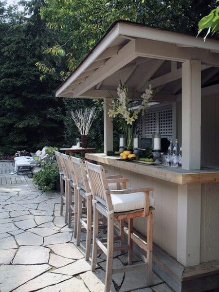 Whitewashed outdoor bar with wooden stools and floral decor on a stone patio.