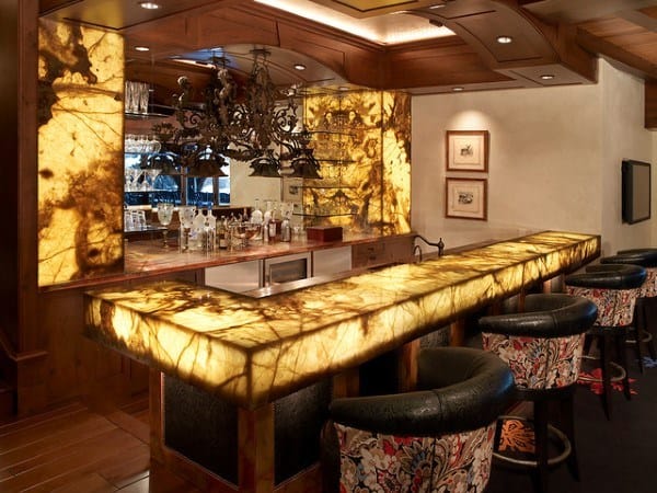 Elegant bar with a glowing amber stone top, ornate chandelier, and chic upholstered seating featuring intricate floral patterns.