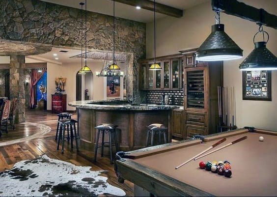 Rustic man cave with pool table, bar area, stone walls, and wooden floors; cozy, warm lighting enhances the ambiance