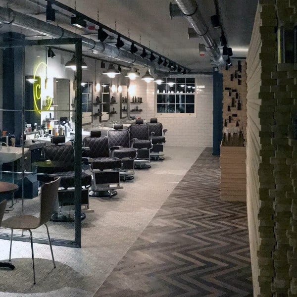 Industrial barbershop with exposed ductwork, quilted chairs, and herringbone flooring.