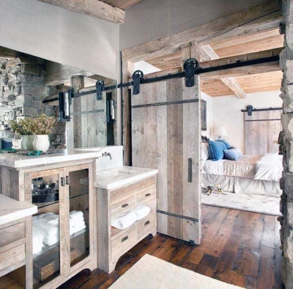 Rustic bathroom with wooden sliding door and vanity, revealing a cozy rustic bedroom