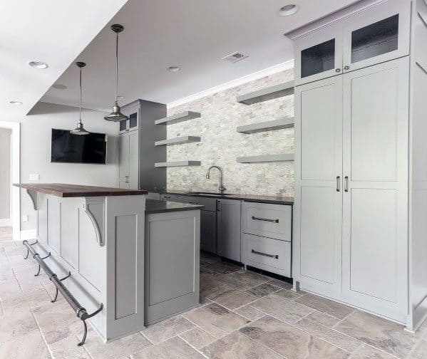 Modern basement bar with light grey cabinetry, stone backsplash, and metal foot rail on tiled floor.