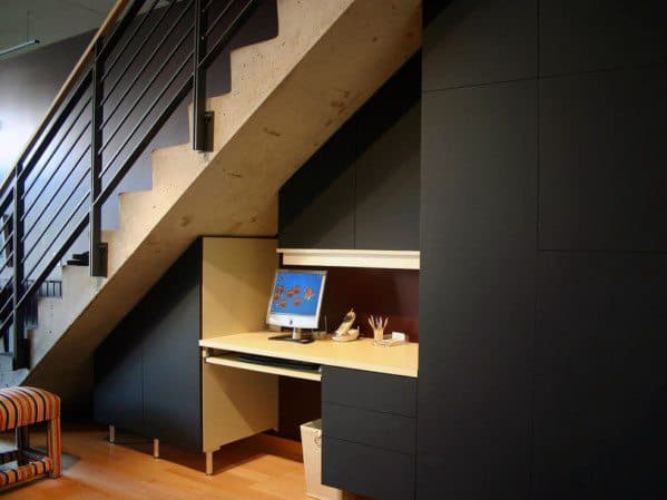 Under-the-stairs office nook with built-in desk, cabinets, and computer in a modern interior design.