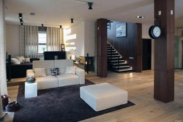 Modern basement with large brown support beams, white furniture, and a staircase backdrop.