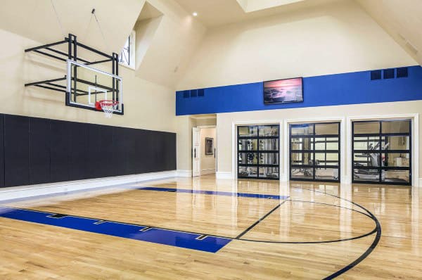 Home indoor basketball court with wooden floors, high ceiling, a mounted hoop, and exercise equipment visible through glass doors