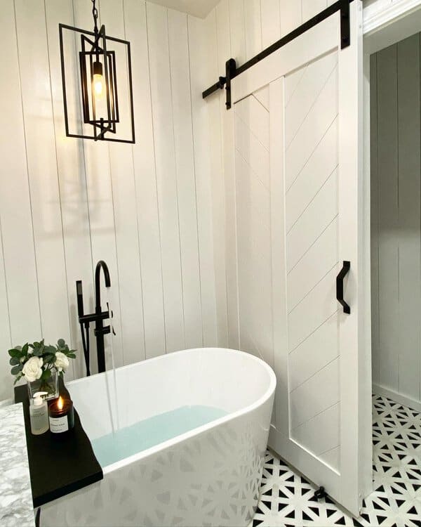 Cage-style black pendant above a white freestanding tub, barn door, and patterned floor in a modern farmhouse bathroom.