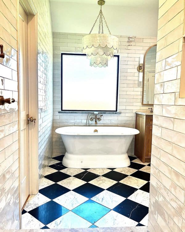 Capiz scallop chandelier above a white freestanding tub with checkerboard floor and subway tile walls in a bright bathroom.
