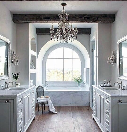 Elegant bathroom with crystal chandelier, arched window, marble bathtub, and exposed wood beams.