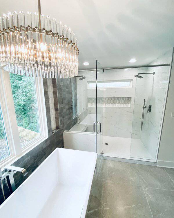 Drum-style chandelier with vertical glass rods above a white rectangular tub in a modern bathroom with large windows and marble accents.
