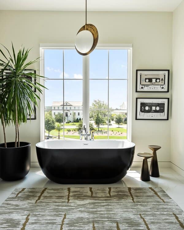 Gold disc pendant above a black freestanding tub, large window, and modern decor in a bright bathroom with a tall houseplant.