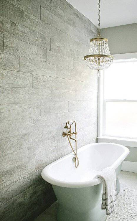 Beaded Empire chandelier above a freestanding tub with wood-look tile walls and brass fixtures in a cozy, rustic bathroom.