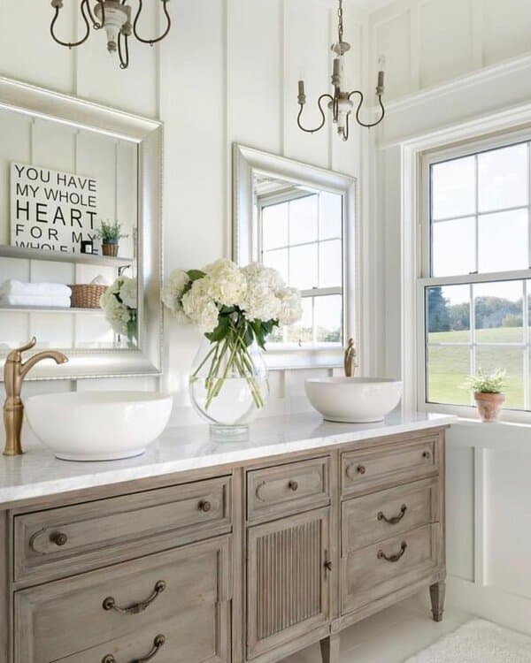 Twin candelabra-style chandeliers above a weathered vanity with marble top in a bright, farmhouse-inspired bathroom.