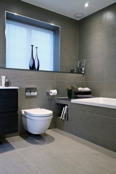Modern bathroom with grey floor and wall tiles, wall-hung toilet, and built-in bathtub with shelving.