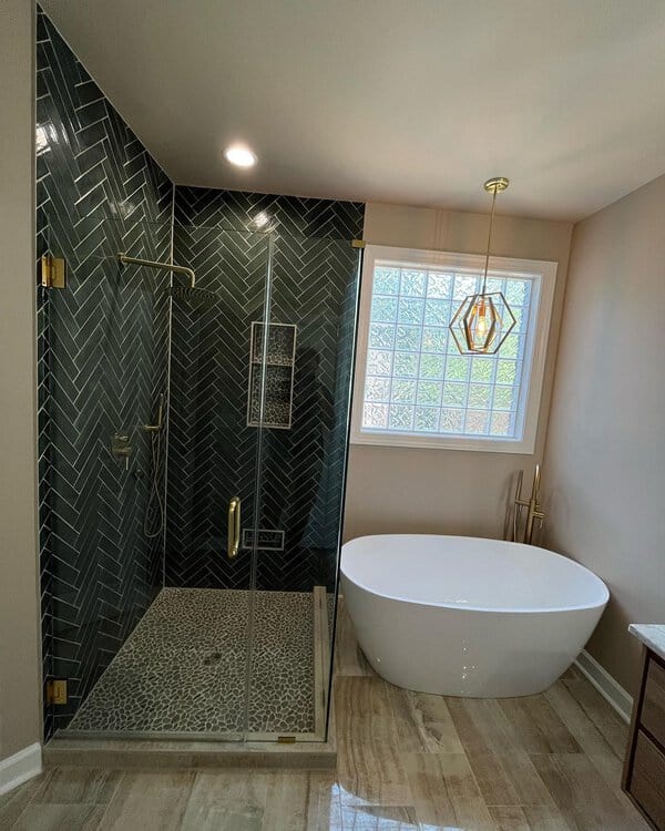 Gold geometric pendant above a white freestanding tub beside a green herringbone-tiled shower with pebble flooring.