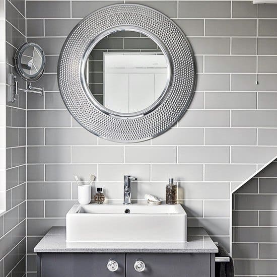 Bathroom with round metallic-framed mirror, gray subway tiles, and white vessel sink.