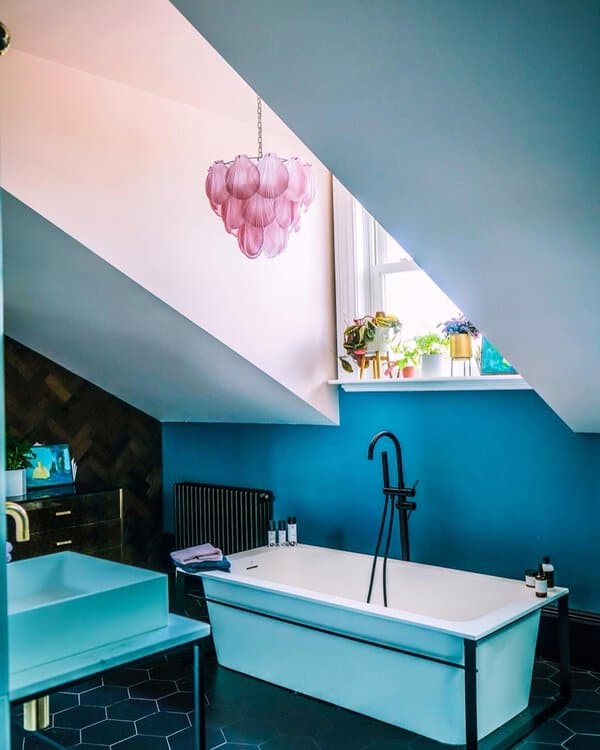 Pink petal chandelier above a white open-frame tub, teal walls, and black hardware in a vibrant attic bathroom.