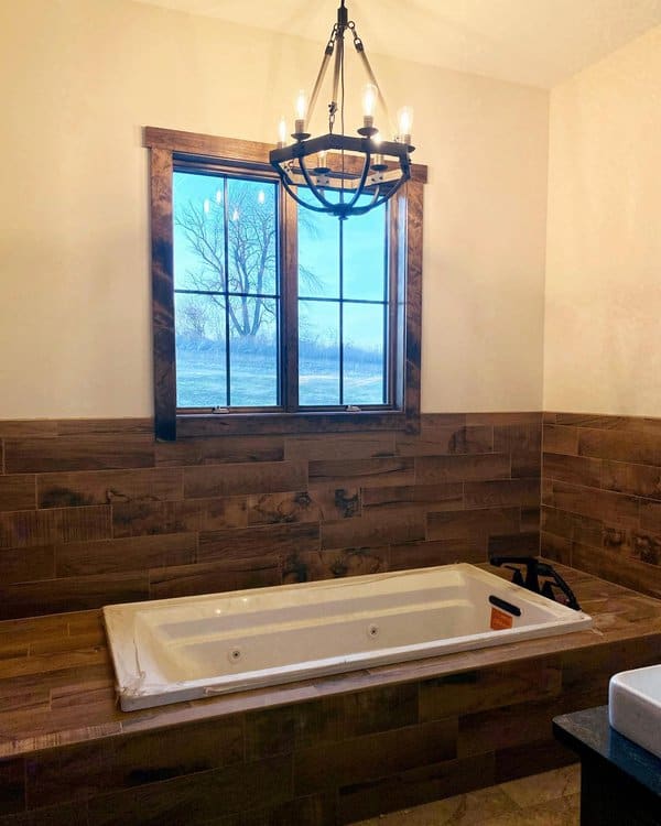 Black industrial chandelier above a built-in jetted tub with wood tile surround and a large window overlooking a rural scene.