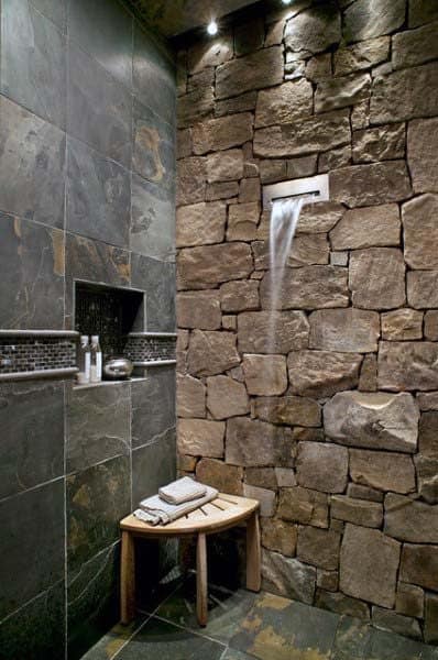 Rustic bathroom with stone walls, a waterfall shower, and a wooden bench draped in towels
