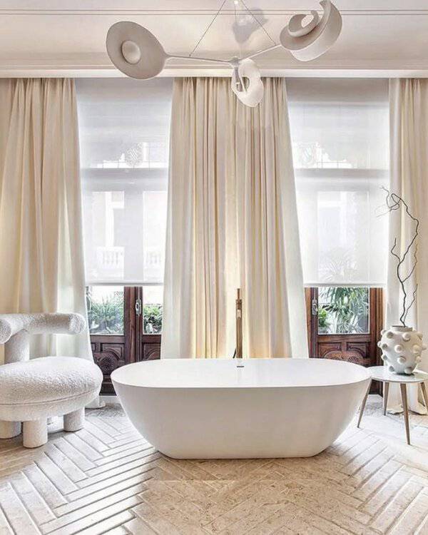 White sculptural chandelier above a freestanding tub, cream drapery, and herringbone flooring in a serene, minimalist bathroom.