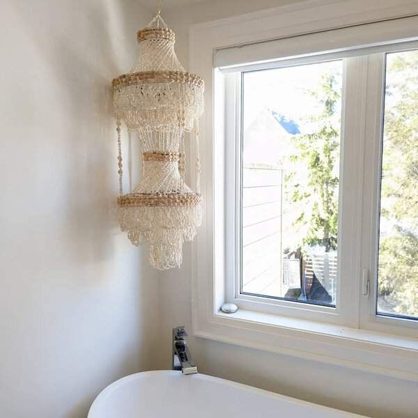 Tiered shell-bead chandelier hanging beside a window in a bright, coastal-inspired bathroom.