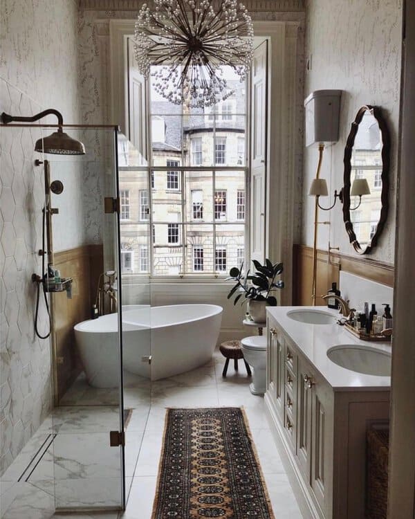 Starburst chandelier above a freestanding tub, tall windows, marble tile, and a vintage runner in a grand, airy bathroom.