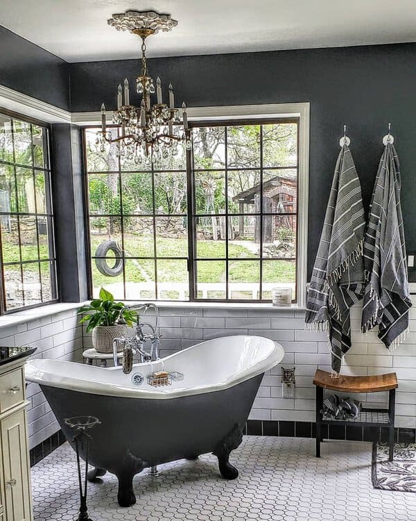 Crystal chandelier above a dark clawfoot tub with white subway tile and big windows for a vintage-meets-modern bathroom.