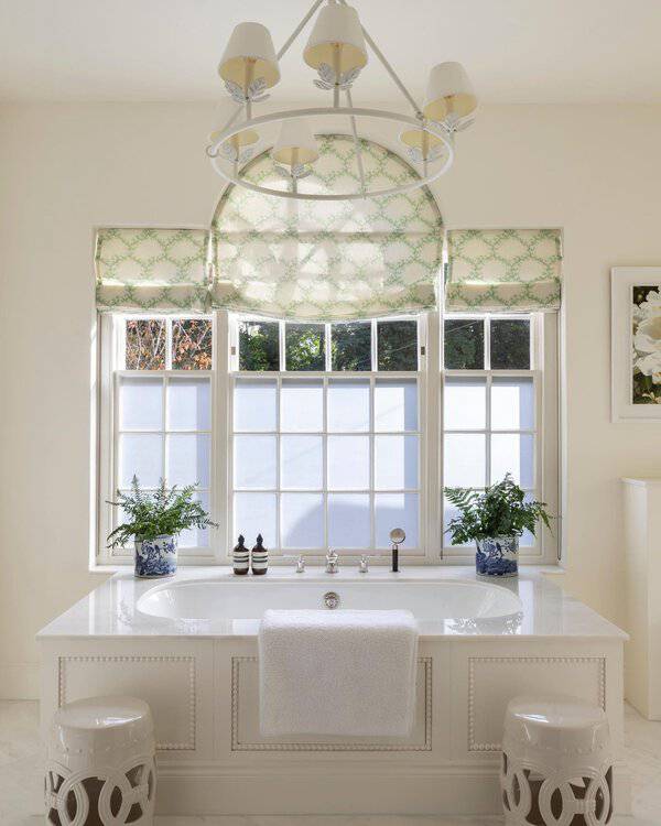 White ring chandelier above a built-in tub, with soft green-patterned shades and blue-and-white planters in a bright bathroom.